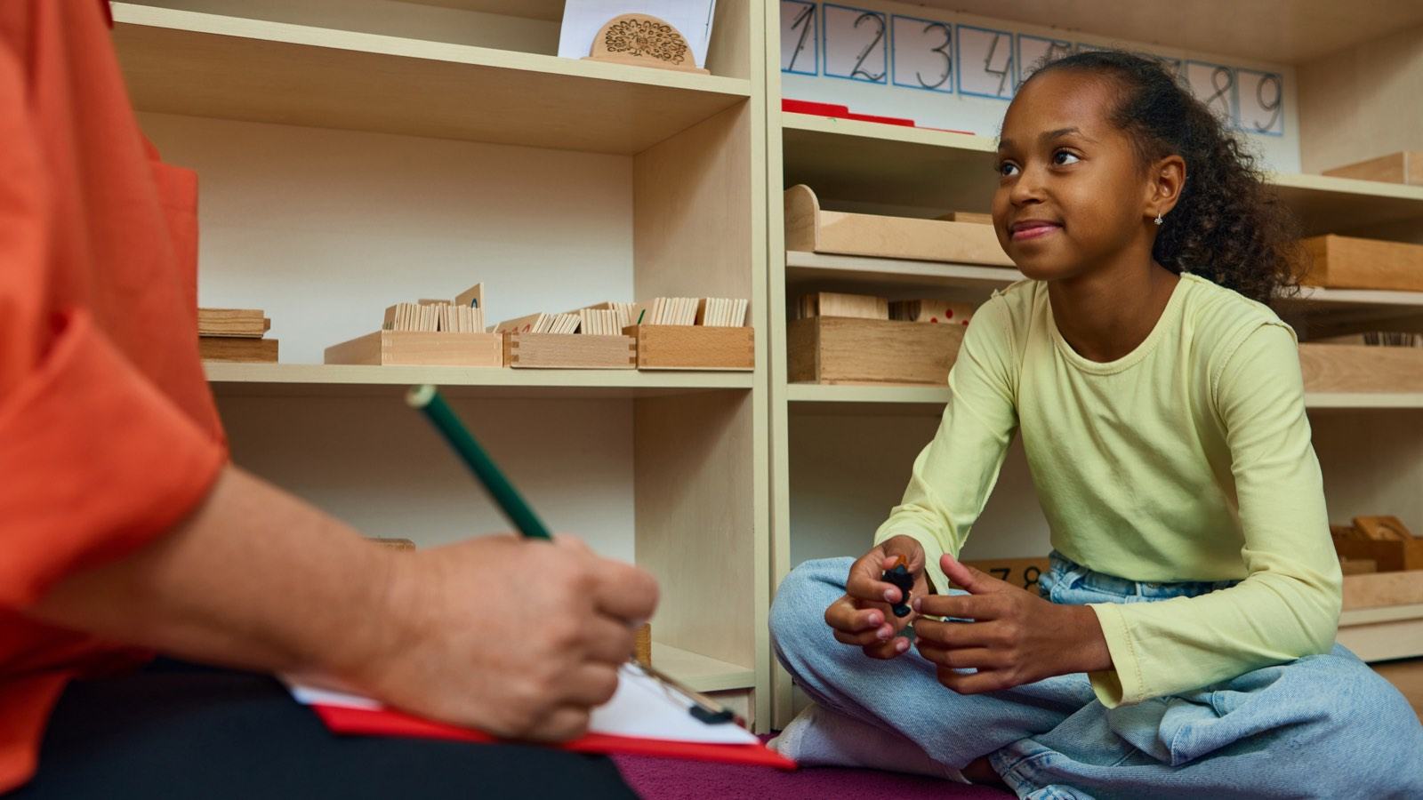 A teacher takes notes on a clipboard while a girl sits cross-legged in a classroom
